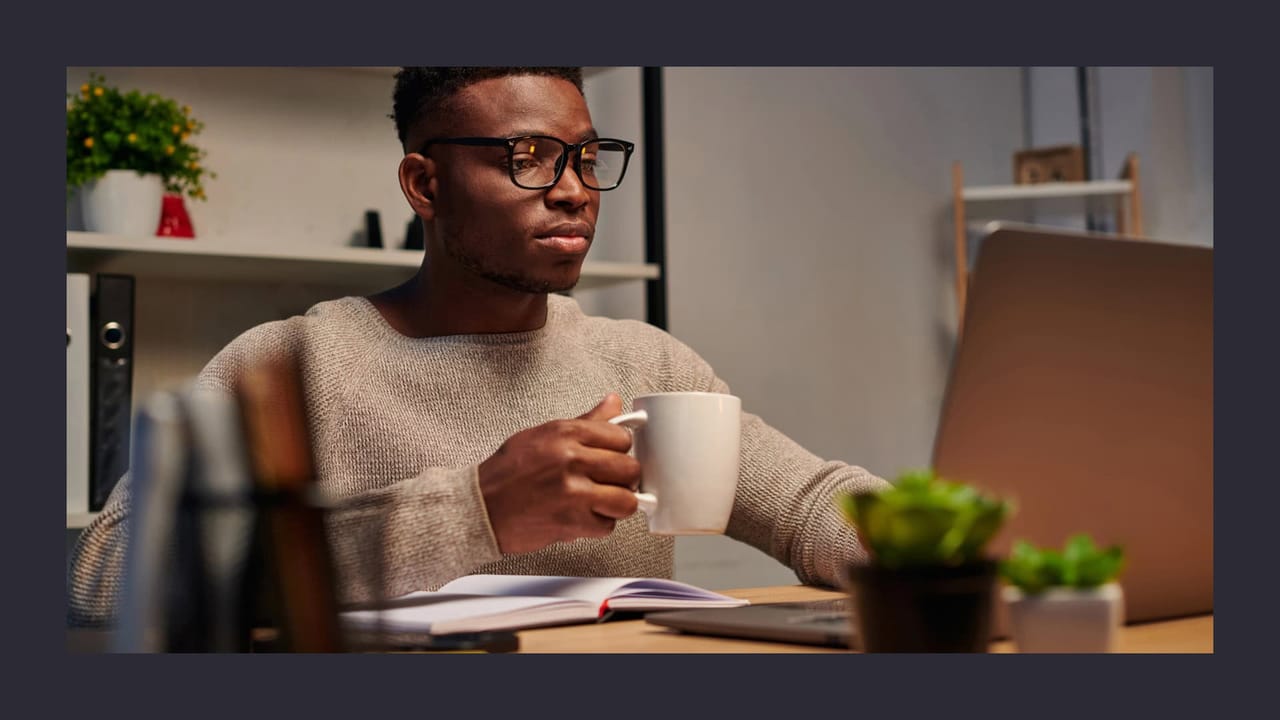 Man with glasses working on laptop, holding coffee mug