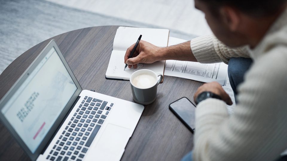 Man writing in notebook alongside laptop and coffee