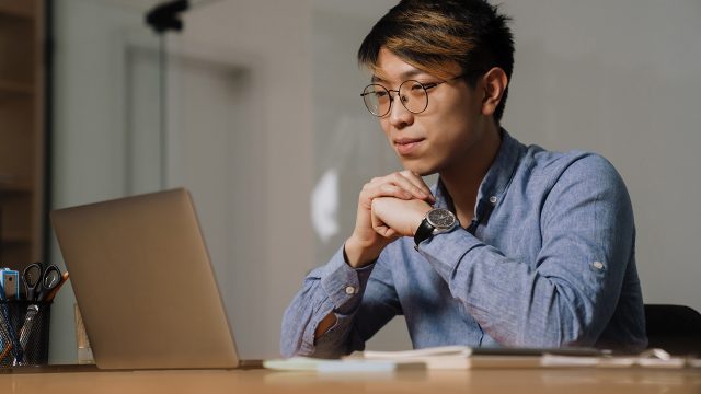 Focused asian guy in glasses researching how much a 5 page website costs on his laptop