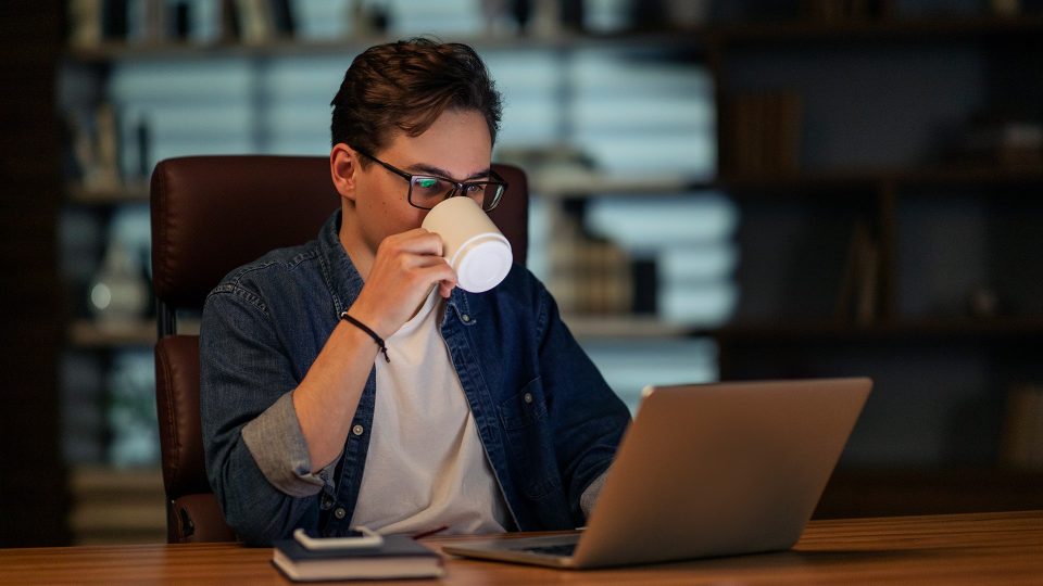 Young man working on his laptop developing a website late in his home office and drinking coffee 