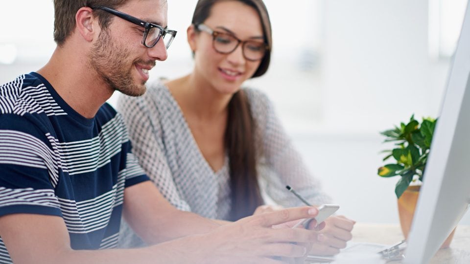 Two coworkers attentively examining a smartphone while seated at a desk in a modern office setting, symbolising the power of mobile computing.